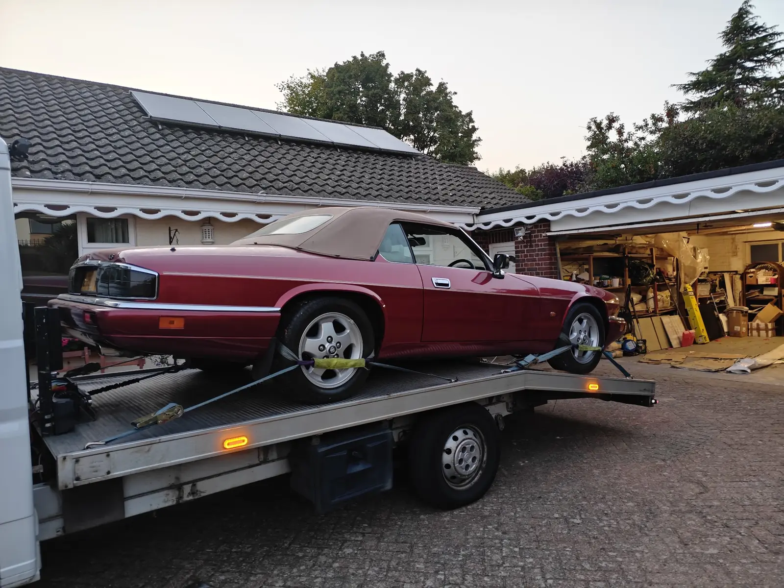 Red Jaguar XJS convertible strapped down on the recovery truck outside a bungalow at dusk